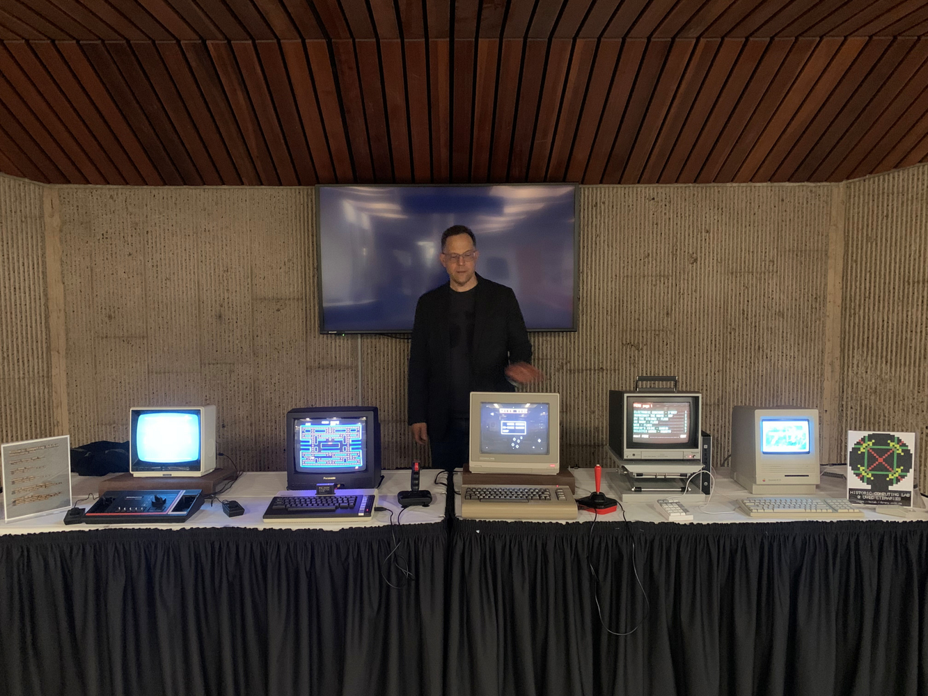 Five computers/consoles mostly dating from the late 1980s on a table, with curator John Durno standing behind
