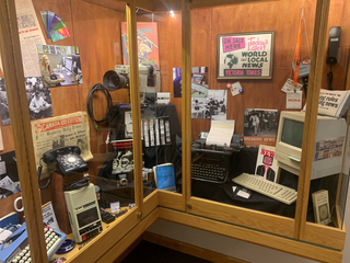 A display cabinet featuring various journalism-related artifacts: video camera, typewriters, tape recorder, rotary dial phone, set against a backdrop of printed ephemera