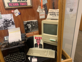 A display cabinet showing a Mac Classic II, an old manual type writer, press passes, and two journalism related books