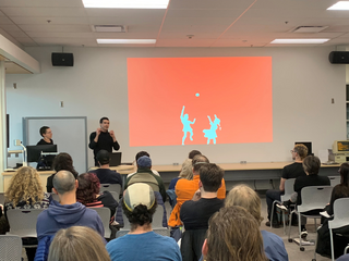 Two presenters dressed in black in front of a classroom audience, with a picture of silhouettes of a boy, girl and a ball projected on the wall