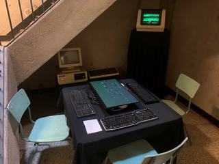 An LCD monitor laid flat on a tabletop, keyboards on 3 sides. An old terminal and beige 1980s computer behind.